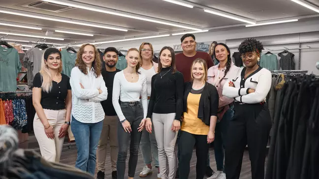 Um grupo de colaboradores encontra-se numa loja, junto a expositores de roupa, a olhar para a câmara.