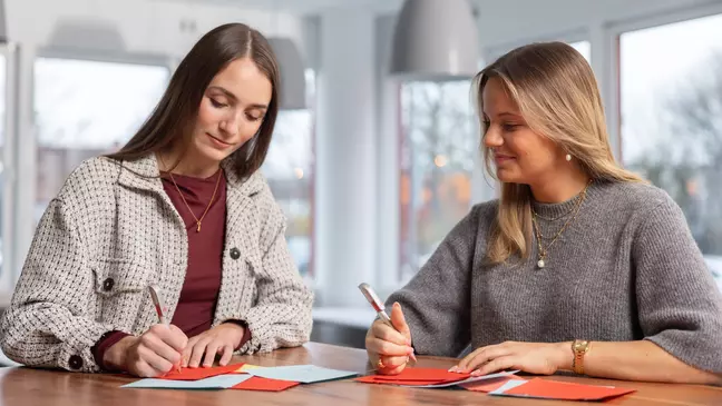 Duas colaboradoras estão de pé junto a uma mesa, com uma caneta na mão, a observar os documentos que se encontram sobre a mesa
