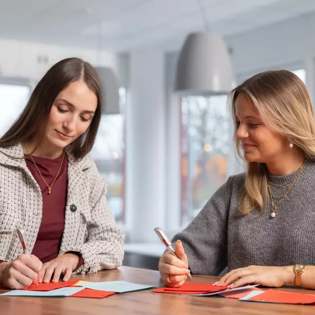 Zwei Mitarbeiterinnen stehen an einem Tisch mit Kugelschreiber in der Hand und schauen auf die Unterlagen die auf dem Tisch liegen