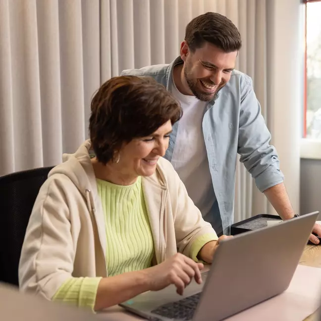 Zwei Mitarbeitende lachen gemeinsam vor einem Laptop in einem modernen Büro
