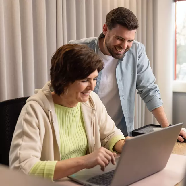 Zwei Mitarbeitende lachen gemeinsam vor einem Laptop in einem modernen Büro