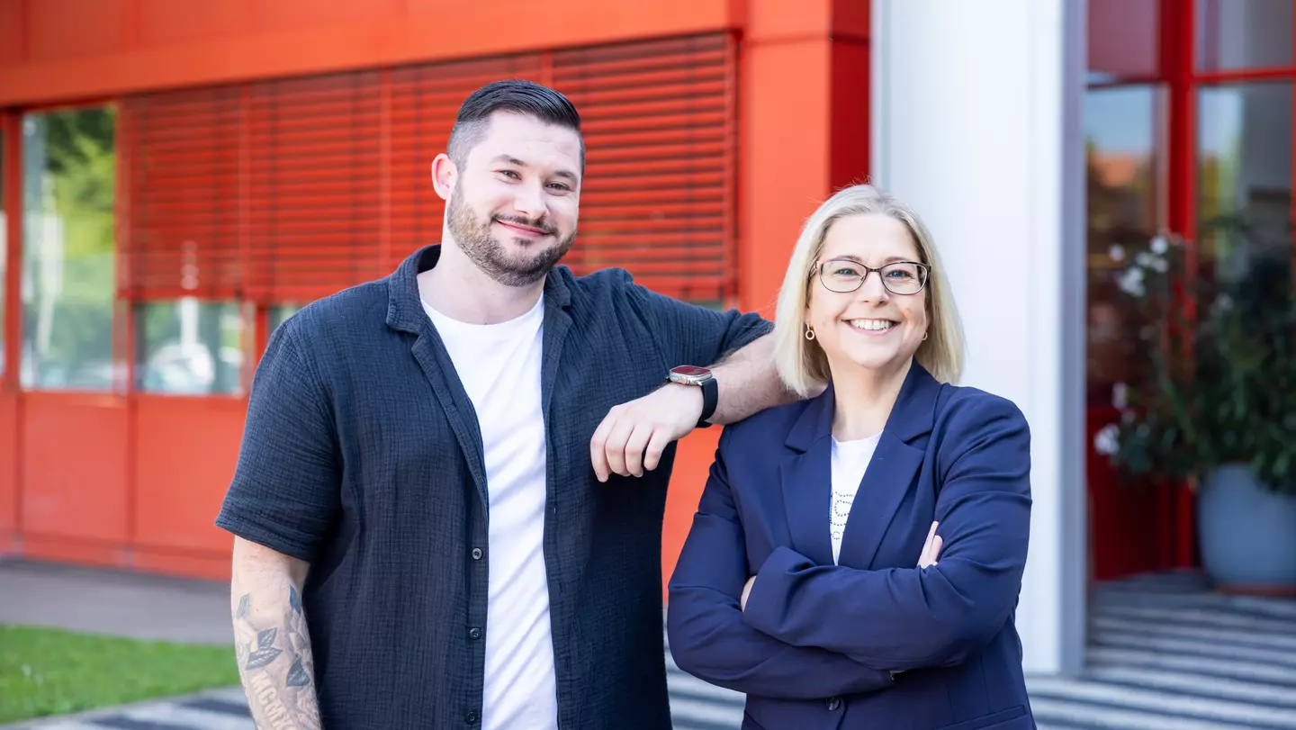 Dois colaboradores encontram-se no exterior de um edifício e sorriem para a câmara.