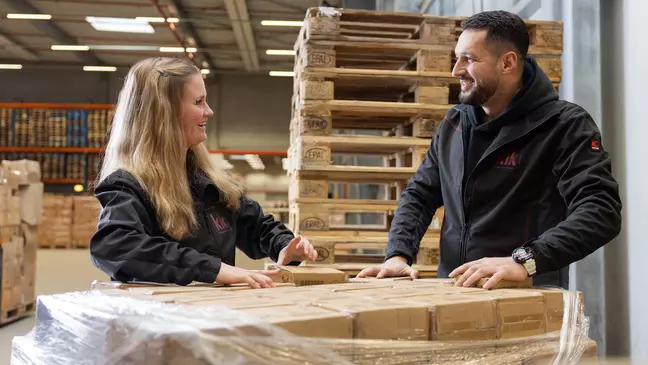 Zwei Mitarbeitende lächeln sich an, sie stehen im Lager und packen Lagerware aus