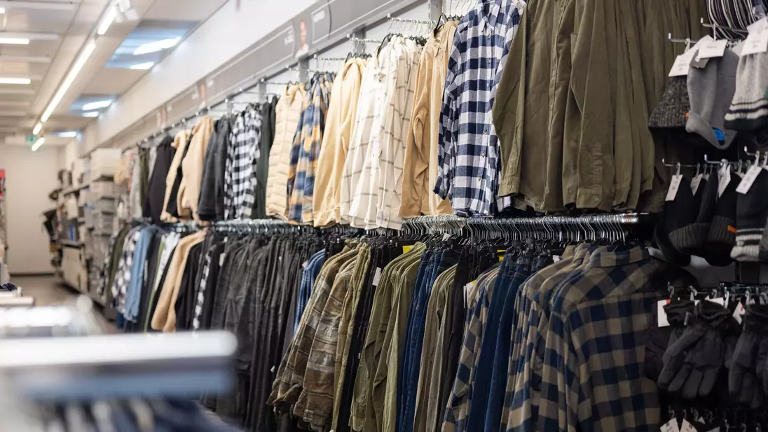 Roupa masculina exposta em varões de roupa junto à parede do fundo de uma loja KiK.