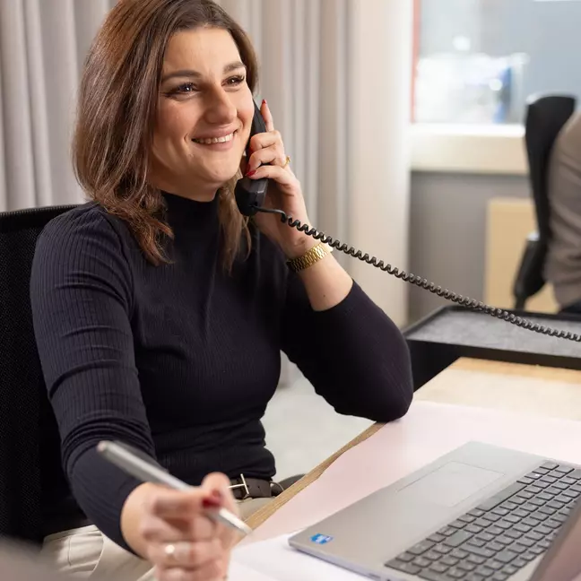 Uma colaboradora está sentada no seu local de trabalho ao telefone e sorri para a câmara
