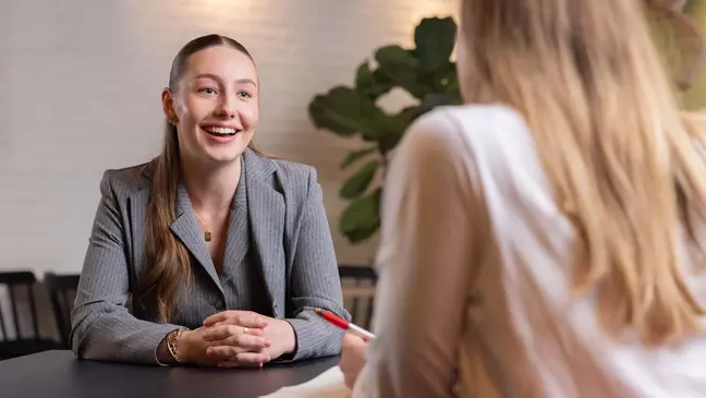 Uma colaboradora fala, sorrindo, com outra colaboradora, junto a uma mesa numa sala