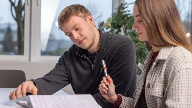 Ein Mann und eine Frau schauen in ein Notizbuch, die Frau hält einen Kugelschreiber, beide lächeln und sitzen in einem modernen Büro
