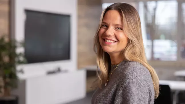 Eine Frau schaut lächelnd in die Kamera, sie steht in einer modernen Räumlichkeit