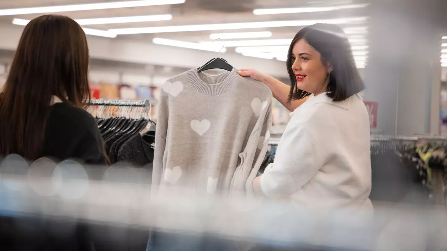 Uma colaboradora apresenta a uma cliente uma camisola cinzenta com um desenho de coração, na loja de roupa.