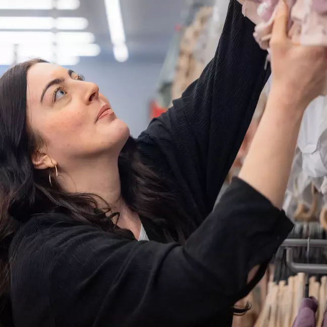 Uma mulher de cabelo escuro estende o braço para alcançar uma prateleira, enquanto procura atentamente algo.
