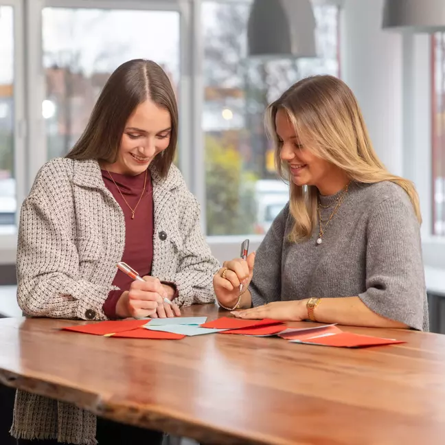 Zwei Mitarbeiterinnen stehen an einem Tisch mit Kugelschreiber in der Hand und schauen auf die Unterlagen die auf dem Tisch liegen