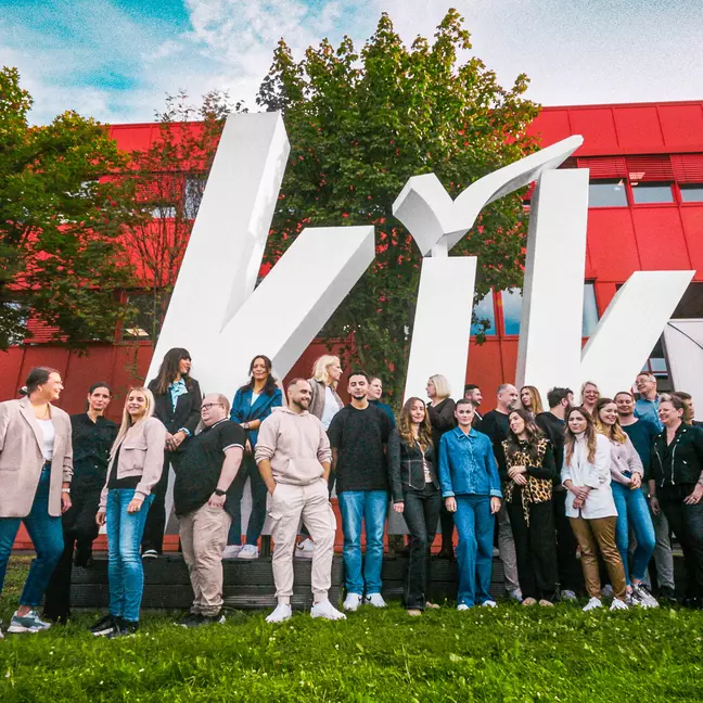 Fotografia de grupo de colaboradores junto à sede europeia da KiK em Bönen, com o grande logótipo da KiK em destaque.