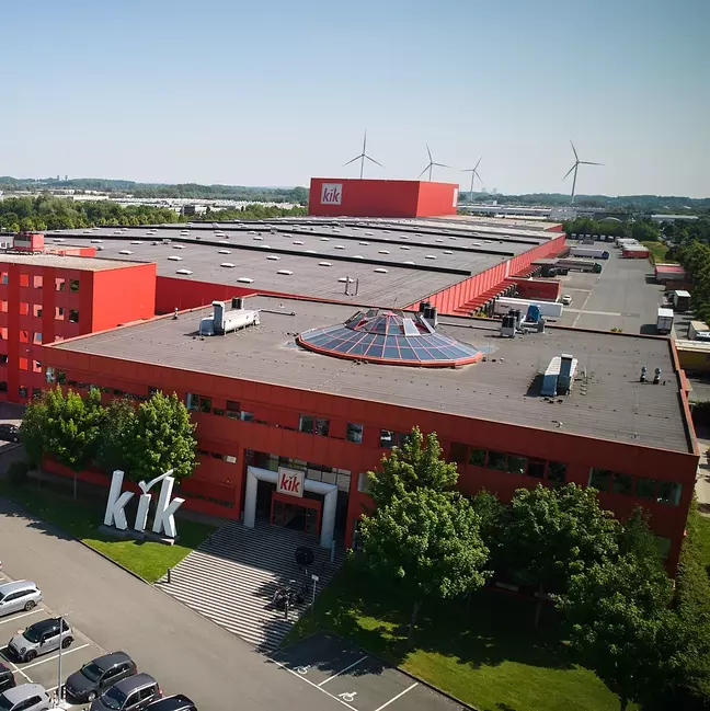 Um grande edifício vermelho da empresa, com o logótipo "KiK", está situado numa paisagem verde.