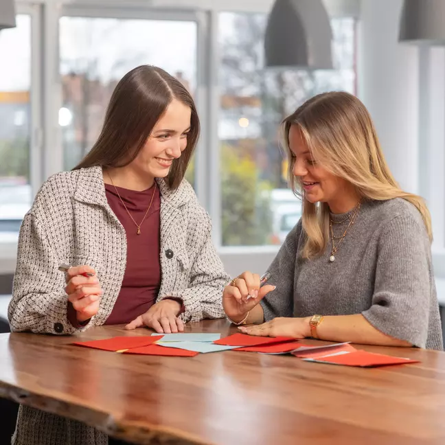 Zwei Mitarbeiterinnen stehen lächelnd an einem Tisch mit Kugelschreiber in der Hand und schauen auf die Unterlagen die auf dem Tisch liegen