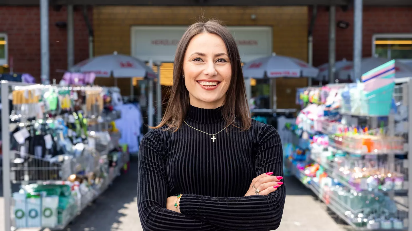 Uma pessoa encontra-se na área exterior em frente à loja, entre expositores de produtos, a sorrir para a câmara.