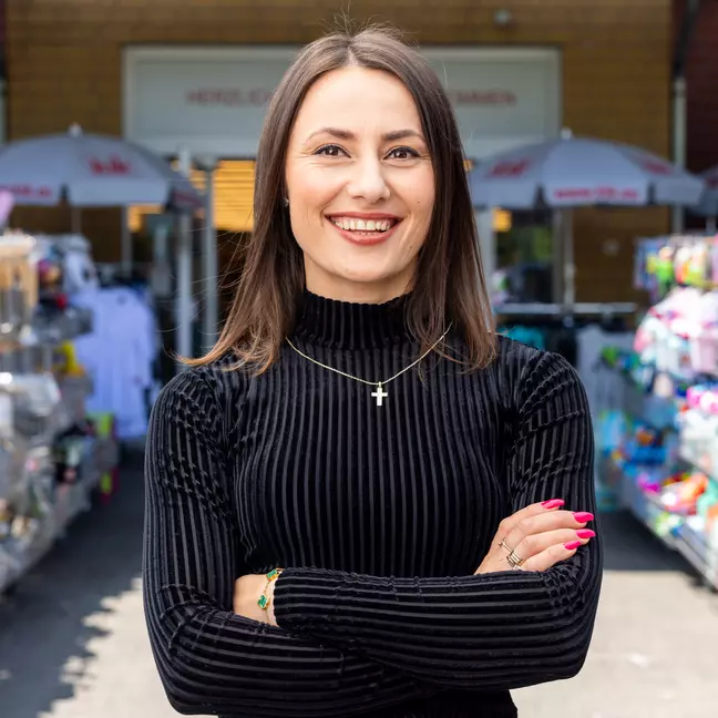Uma pessoa está na zona exterior, em frente à loja, entre as prateleiras de produtos, e olha sorrindo para a câmara.