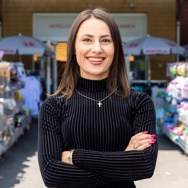 Uma colaboradora encontra-se no exterior, em frente a uma loja com expositores de mercadoria, e olha sorridente para a câmara.