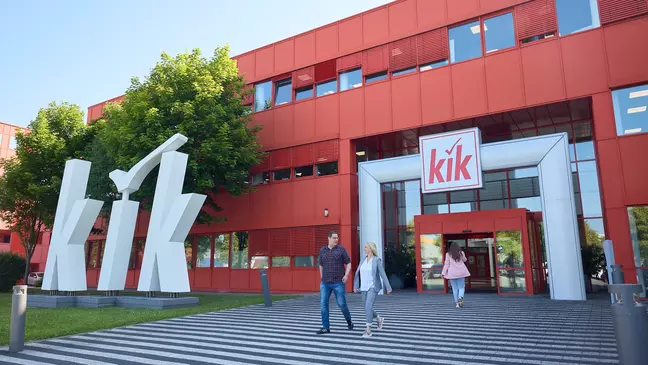 Edifício vermelho da KiK, com um grande logótipo branco na entrada, enquanto várias pessoas se dirigem para o seu interior.
