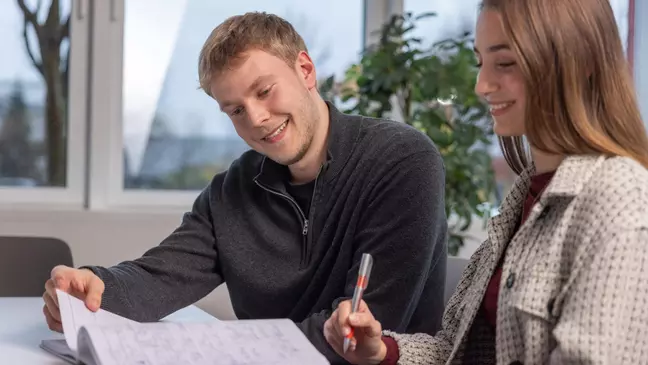 Ein Mann und eine Frau schauen in ein Notizbuch, die Frau hält einen Kugelschreiber, beide lächeln und sitzen in einem modernen Büro