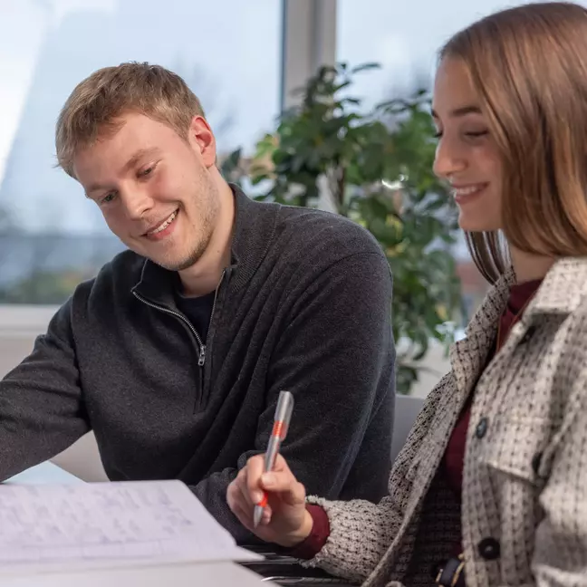 Ein Mann und eine Frau schauen in ein Notizbuch, die Frau hält einen Kugelschreiber, beide lächeln und sitzen in einem modernen Büro