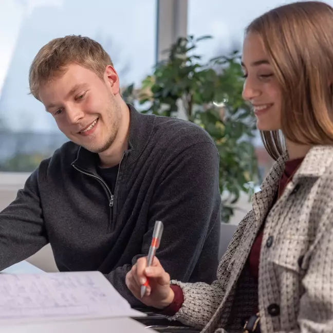 Ein Mann und eine Frau schauen in ein Notizbuch, die Frau hält einen Kugelschreiber, beide lächeln und sitzen in einem modernen Büro